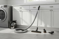 a basic central vacuum attachment kit: hose, floor brush, telescopic wand, upholstery tool, dusting brush, crevice tool, wall hose hanger and storage bag shown in a bright laundry room with tile floors and white cabinets