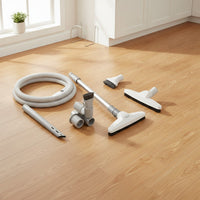 a central vacuum attachment kit for bare floors: deluxe hose, hard floor brush, telescopic wand, upholstery tool, dusting brush, crevice tool, tool caddy and wall hose hanger shown in a sunlit kitchen with wide-plank wood floors and a window