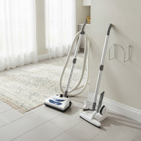 the Johnny Vac PN11 electric central vacuum attachment kit: PN11 power nozzle, dual-voltage hose, telescopic wands, floor brush, upholstery tool, dusting brush, crevice tool, tool caddy and wall hose hanger shown in a bright open-plan kitchen and dining area with pale tile floors and a runner rug